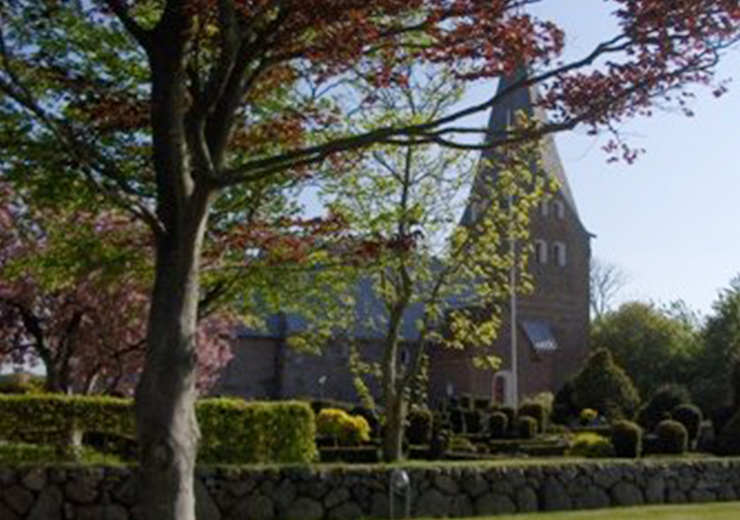 Billede af en kirke med spidst tårn set bag træer i forgrunden.